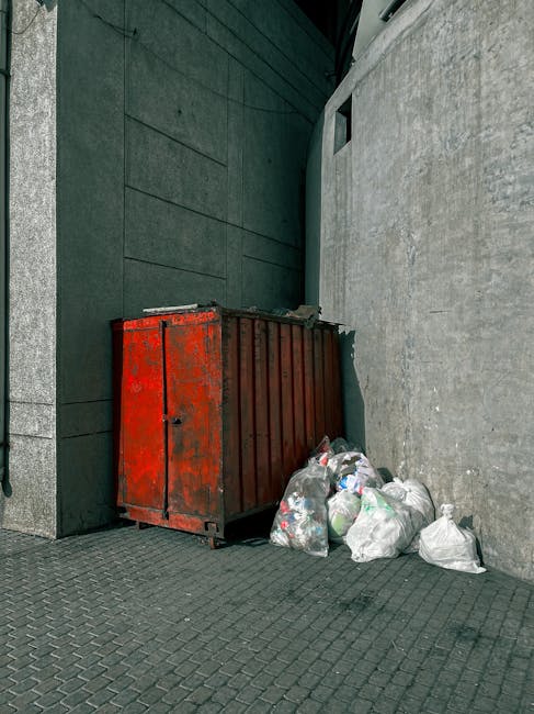 A red metal waste container with visible rust and chipped paint is situated against a dark green wall on the left and a concrete building wall on the right, on a textured paving surface. Around the base of the container, several tied plastic bags filled with mixed rubbish are piled, with some bags partially leaning against it. The scene appears to be an urban exterior alleyway or side street, with minimal natural light contributing to a subdued atmosphere. The overall environment suggests an area designated for non-official waste collection, consistent with private rubbish removal or on-site clearance services often provided by companies like Waste Disposal Roehampton. The composition emphasizes the practical aspect of waste storage and disposal, capturing the typical appearance of discarded rubbish awaiting collection outside a commercial or residential property. The visible details highlight the material and colour of the container, the cluttered arrangement of the rubbish bags, and the surrounding building materials, offering a clear view of a typical waste management setup in an urban setting.
