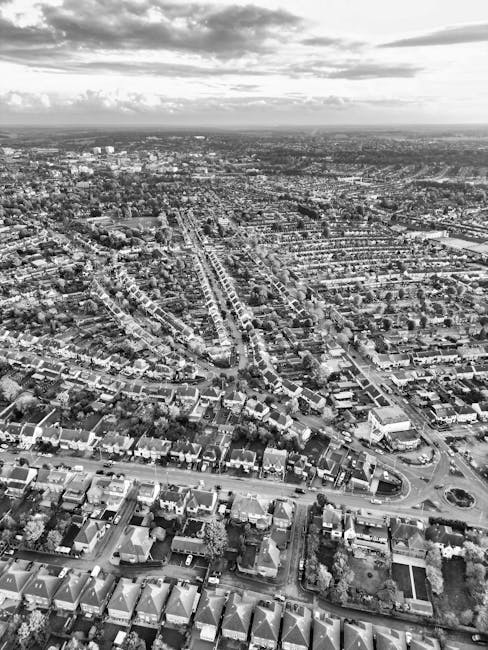 Aerial black-and-white photograph of a densely populated residential area featuring numerous terraced houses with uniform pitched roofs and small backyard gardens. The houses are closely arranged, with street layouts including curved and straight roads, some with roundabouts visible in the foreground. Trees and greenery line parts of the streets and gardens, providing natural contrast against the brick and tile finishes of the buildings. In the distance, the landscape extends towards the horizon, with larger buildings and open spaces visible under a partly cloudy sky. The environment appears calm, with no visible vehicles or pedestrians. The scene presents an overview typical of urban neighbourhoods, where private waste collection and rubbish removal services, such as those offered by Waste Disposal Roehampton, could be relevant for managing household waste in this area. The lighting suggests a diffuse, overcast sky, with no harsh shadows or direct sunlight.