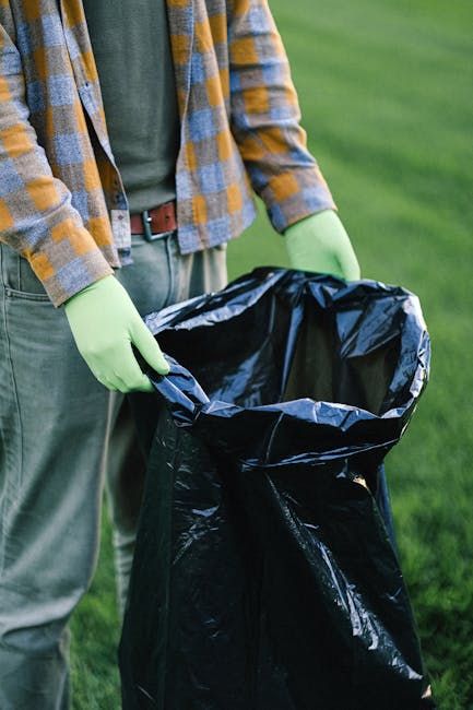 The image shows a person standing outdoors on a grassy area, holding open a large black rubbish bag lined with a silver inner layer. The individual is wearing a long-sleeved plaid shirt in shades of yellow, gray, and blue over a green top, with a brown belt visible around the waist. They are also wearing light green disposable gloves, suggesting they are engaged in waste collection or disposal activities. The background consists of a well-maintained green lawn, and the lighting indicates daytime with natural light illuminating the scene. The black rubbish bag appears to be empty or nearly so, prepared for waste collection, which may relate to private waste handling or on-site clearance processes often performed by waste disposal services like Waste Disposal Roehampton. The scene emphasizes careful handling of waste materials, supporting the context of rubbish removal or alternative waste management methods rather than municipal collection, and highlights the importance of proper disposal practices in environmental upkeep.